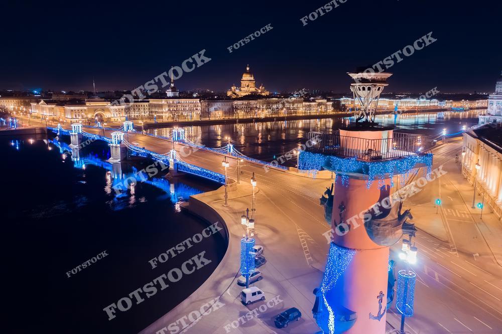 На фотографии изображена Стрелка Васильевского острова, украшенная для праздничного зимнего сезона. Ростральные колонны, украшенные гирляндами, и Дворцовый мост с подсветкой придают ночному Петербургу особое очарование. На заднем плане величественно возвышается Исаакиевский собор, создавая архитектурный акцент. Огни города отражаются в водах Невы, подчёркивая красоту ночного Петербурга и его праздничную атмосферу.